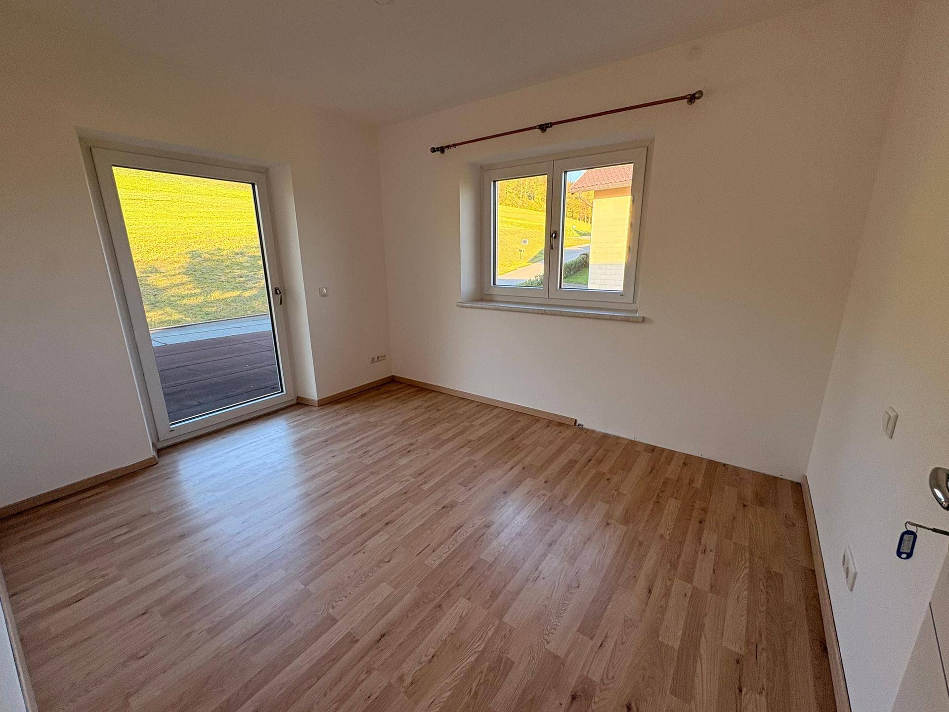 Helles, leeres Zimmer mit Laminatboden, weißen Wänden und Zugang zu einem Außenbereich.