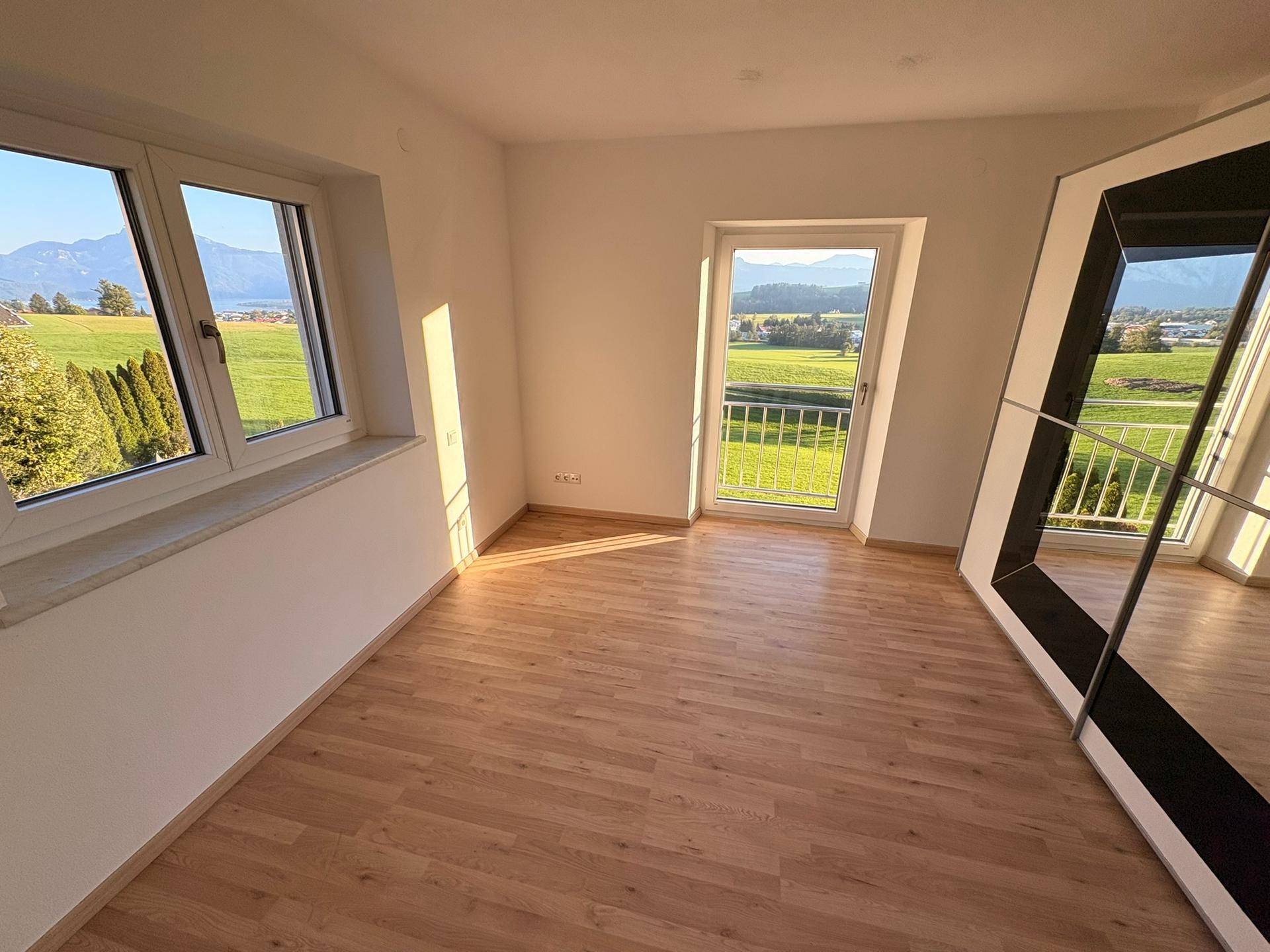 Helles Zimmer mit Laminatboden, großem Fenster und Zugang zu einem französischen Balkon.