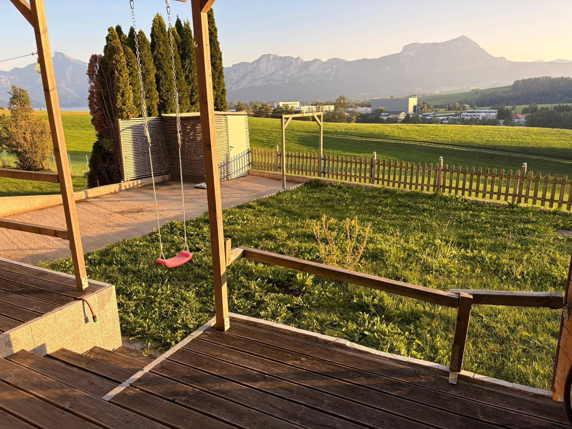 Gepflegter Gartenbereich mit Schaukel und Holzterrasse, umgeben von grünen Wiesen und Bergblick.