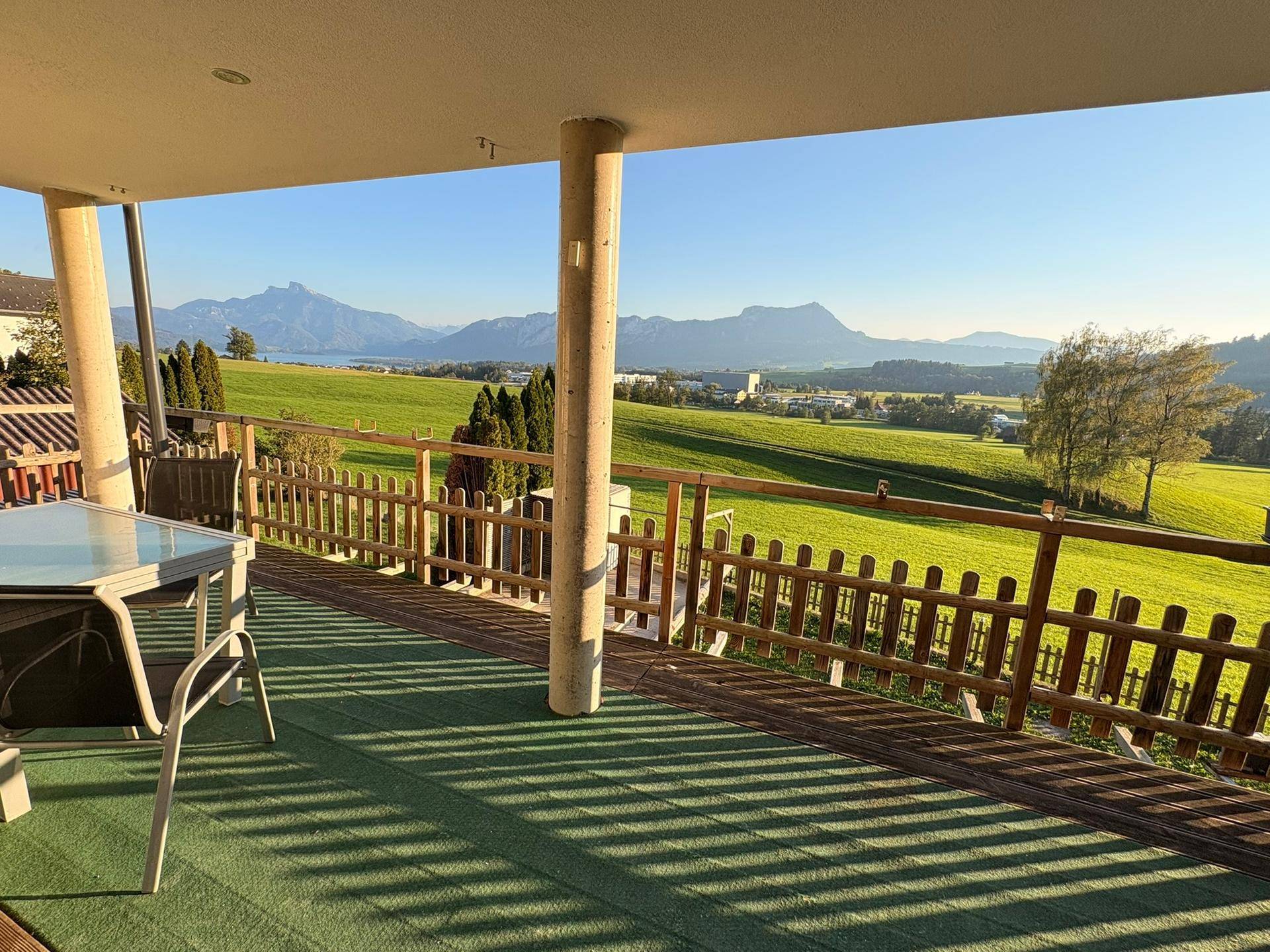 Überdachte Terrasse mit Gartenmöbeln und weitem Blick auf grüne Landschaft, See und Berge.