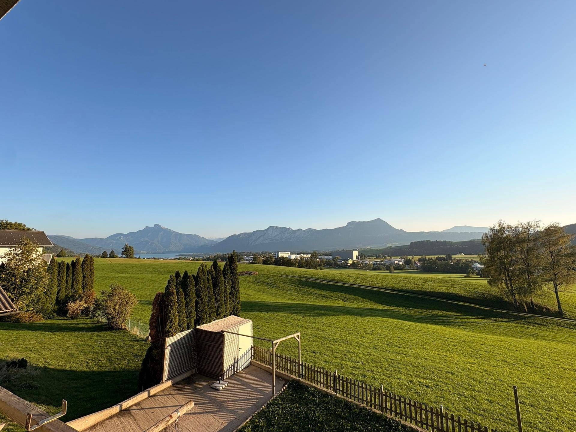 Weitläufiger Ausblick über grüne Wiesen, einen See und die umliegende Berglandschaft.