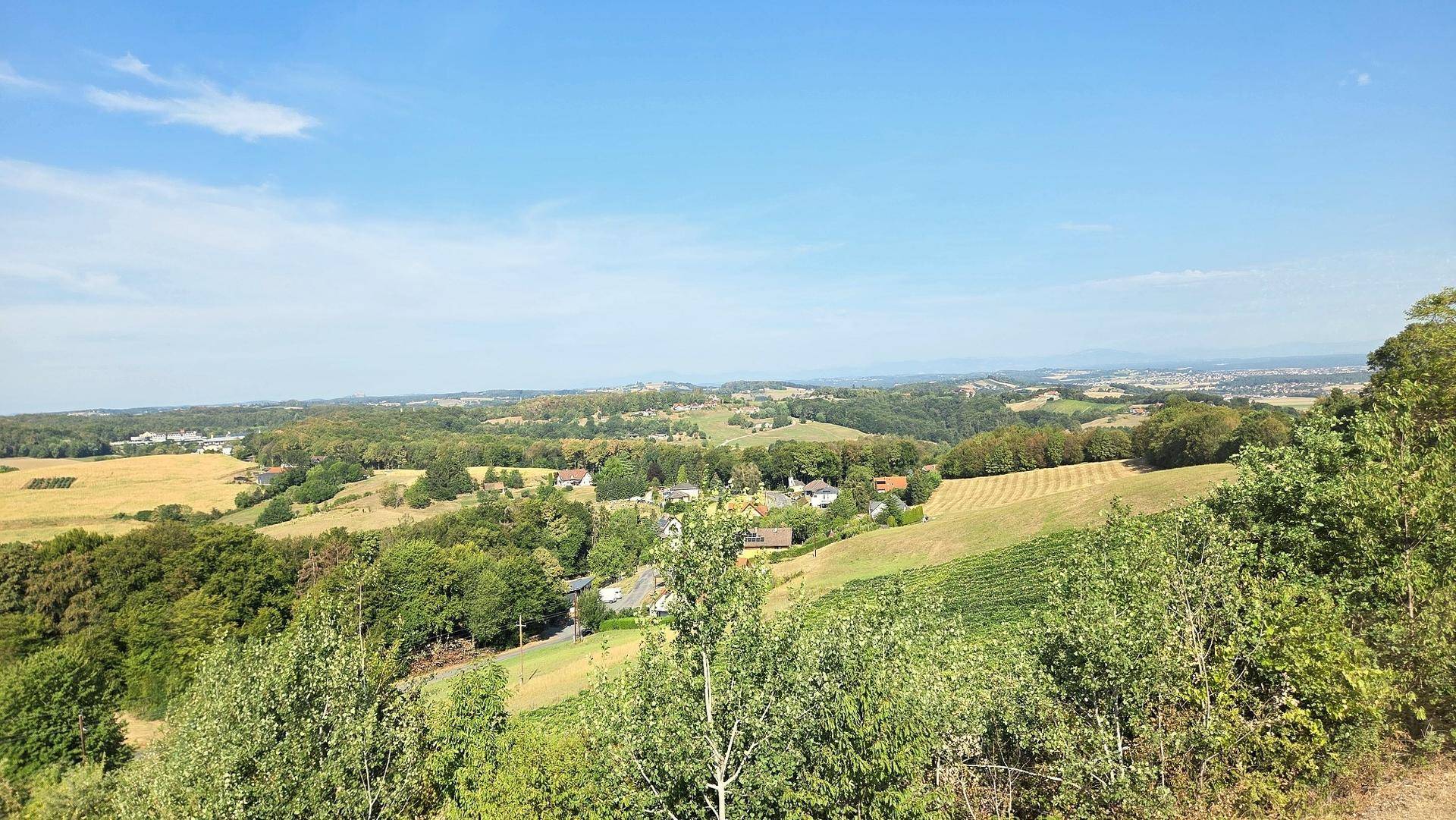 Weitläufige Landschaftsaufnahme mit grünen Hügeln, Feldern und vereinzelten Häusern unter blauem Himmel.
