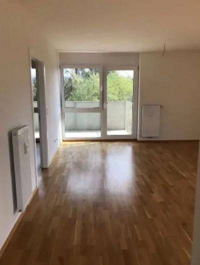 Heller Wohnbereich mit Holzboden und Zugang zu einem Balkon, umgeben von grünen Bäumen.