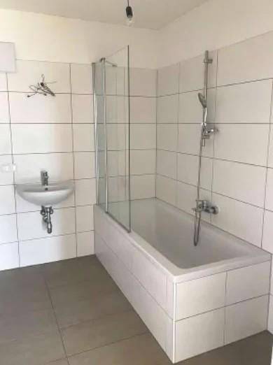 Helles Badezimmer mit weißen Fliesen, Badewanne mit Duschwand und Waschbecken.