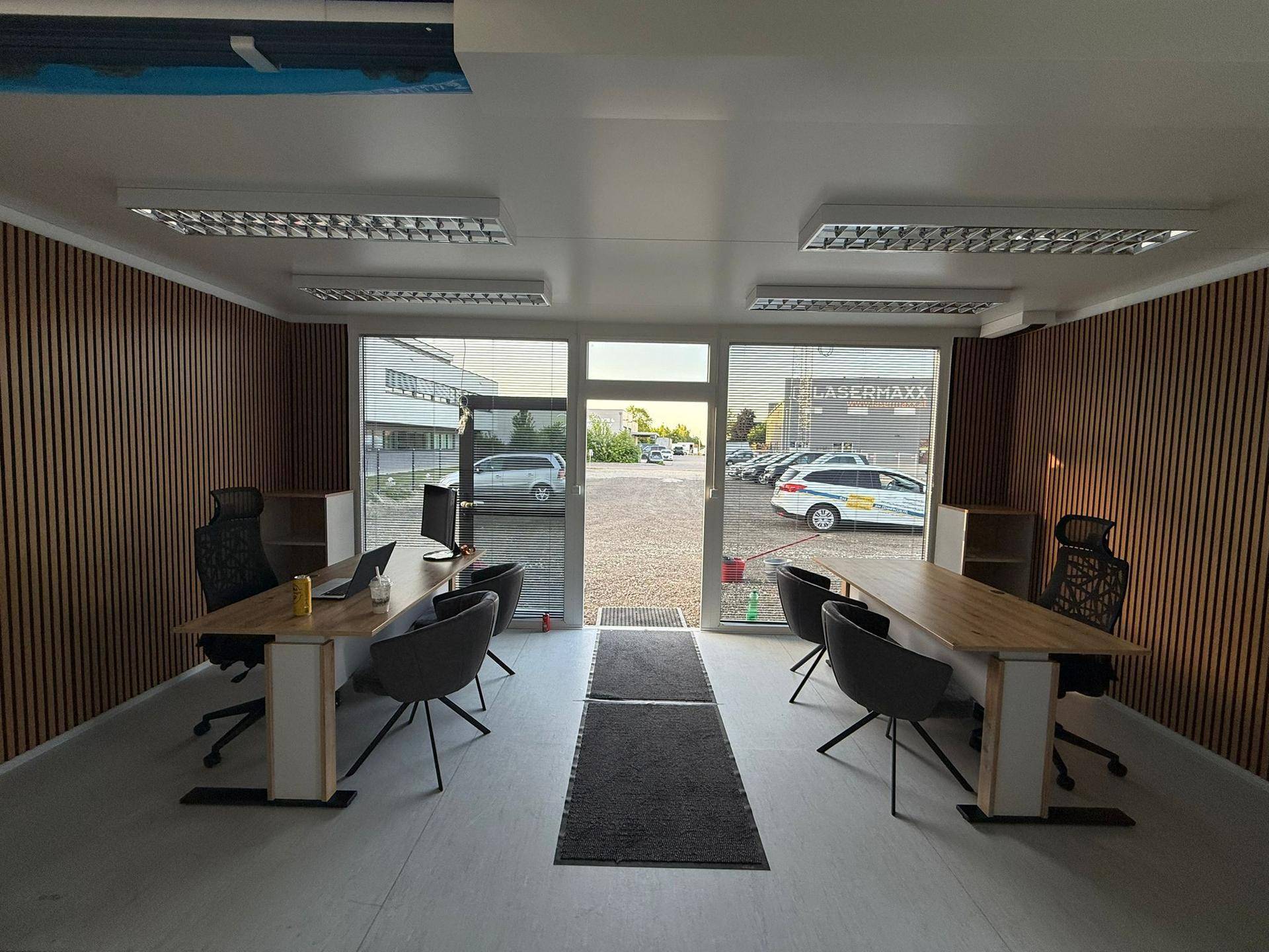 Geräumiges Büro mit zwei Arbeitsplätzen, Holzwandverkleidung und großen Fenstern mit viel Tageslicht.