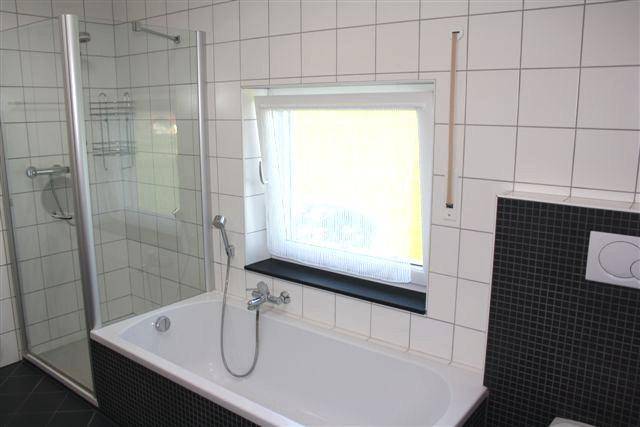 Badezimmer mit Badewanne, integrierter Dusche, Fenster und weißen Wandfliesen.