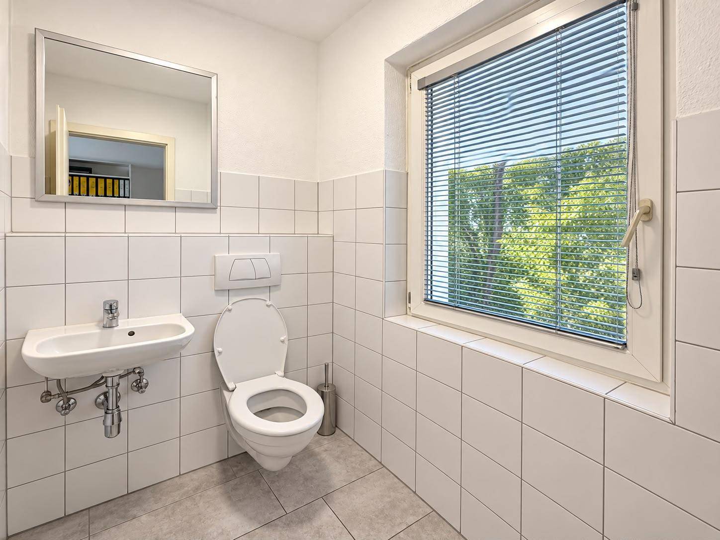 Helles Gäste-WC mit weißen Fliesen, wandhängendem WC, Waschbecken und einem Fenster mit Jalousie.