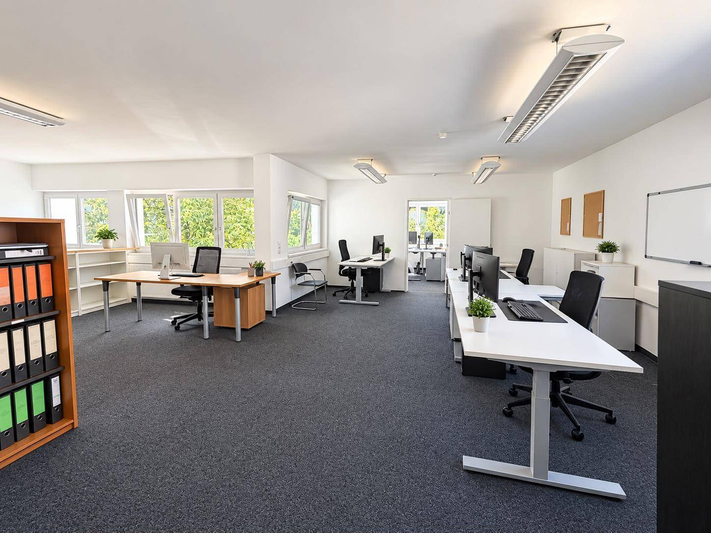 Großes, offenes Büro mit mehreren Arbeitsplätzen, Computern und viel Tageslicht durch die Fenster.