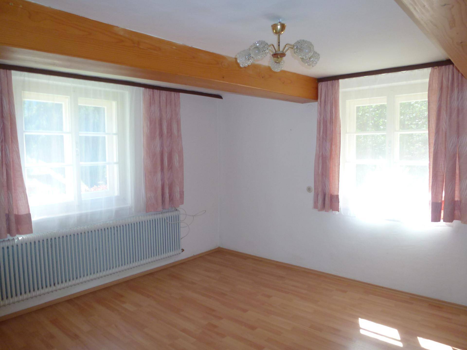 Helles Zimmer mit sichtbaren Holzbalken, Laminatboden und zwei Fenstern mit Vorhängen.