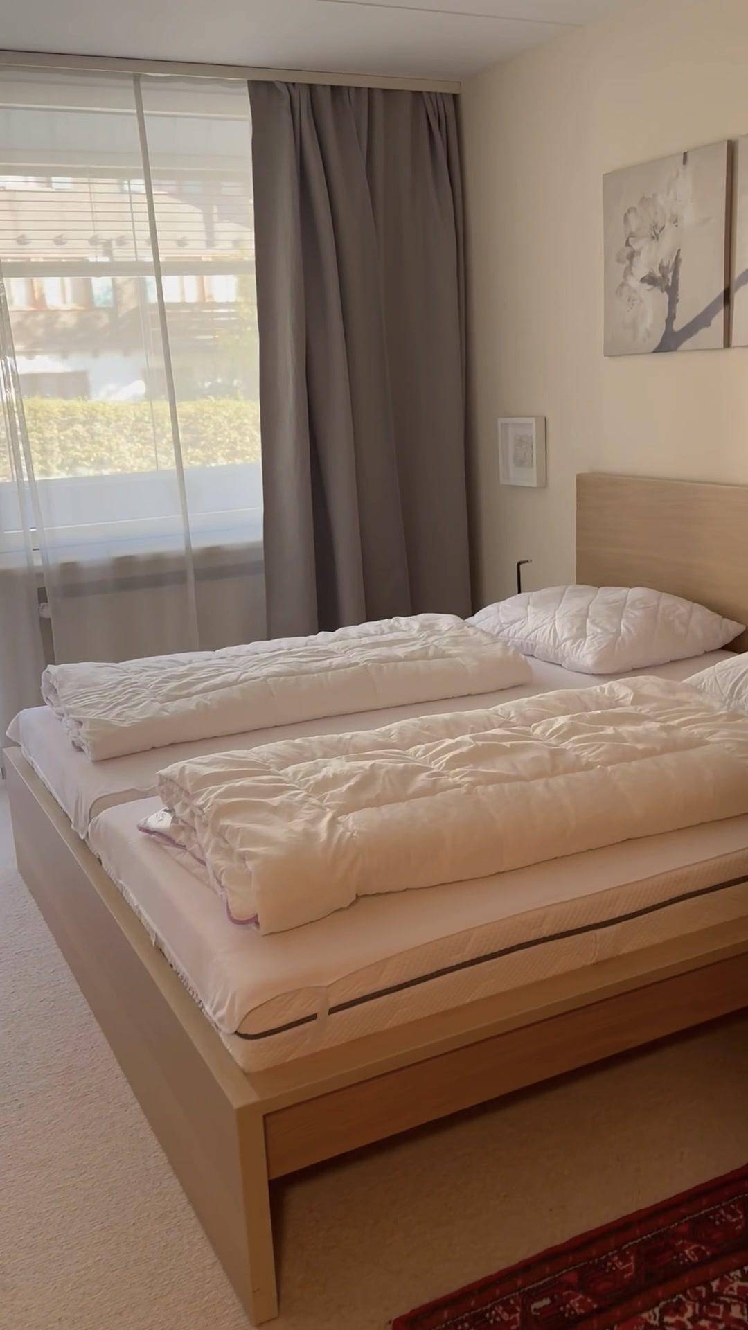 Helles Schlafzimmer mit Doppelbett, Holzbettrahmen und großen Fenstern mit Vorhängen.