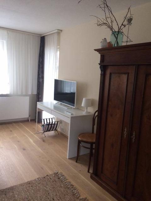 Helles Zimmer mit Holzboden, weißem Schreibtisch, Fernseher und großem Holzschrank für vielseitige Nutzung.