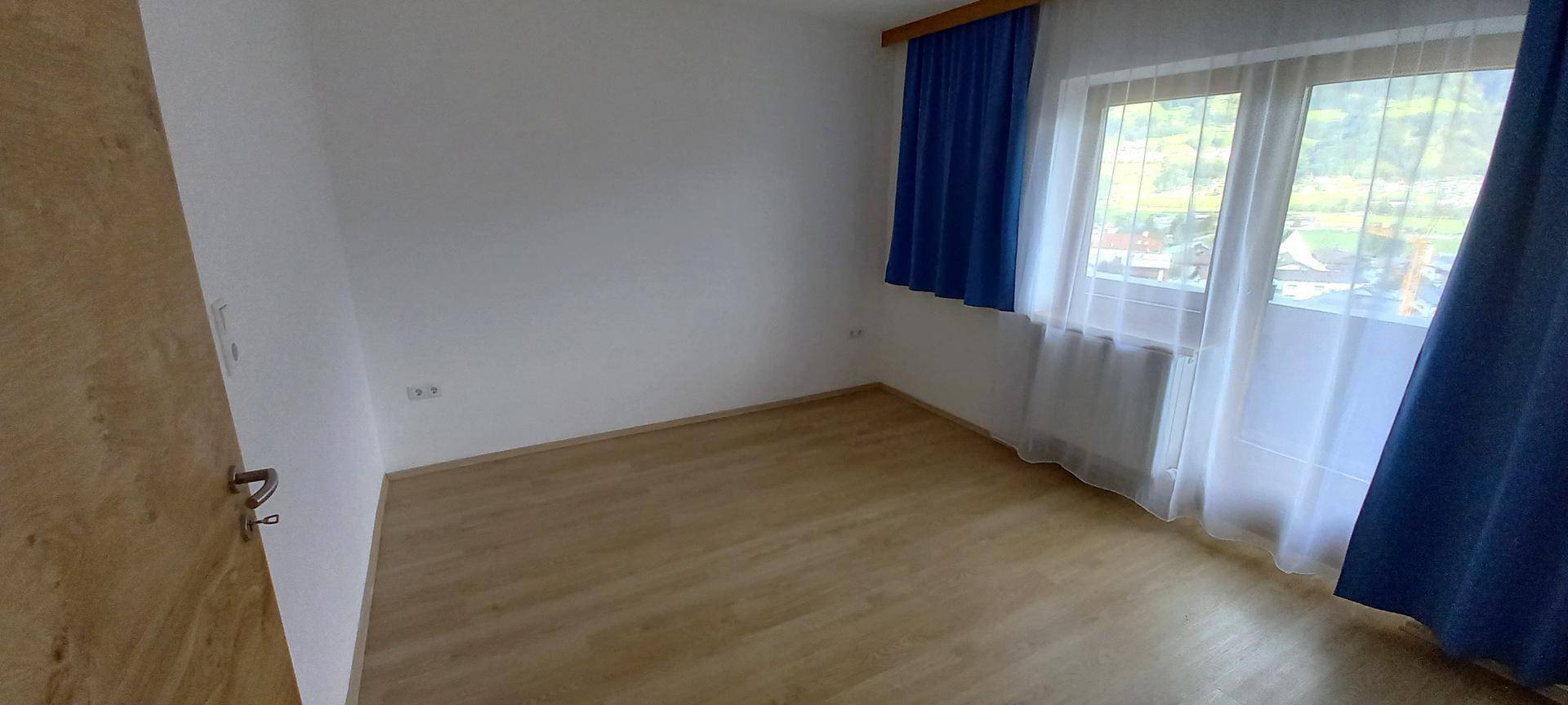 Gemütliches Zimmer mit Holzboden, blauen Vorhängen und einem schönen Blick ins Grüne.