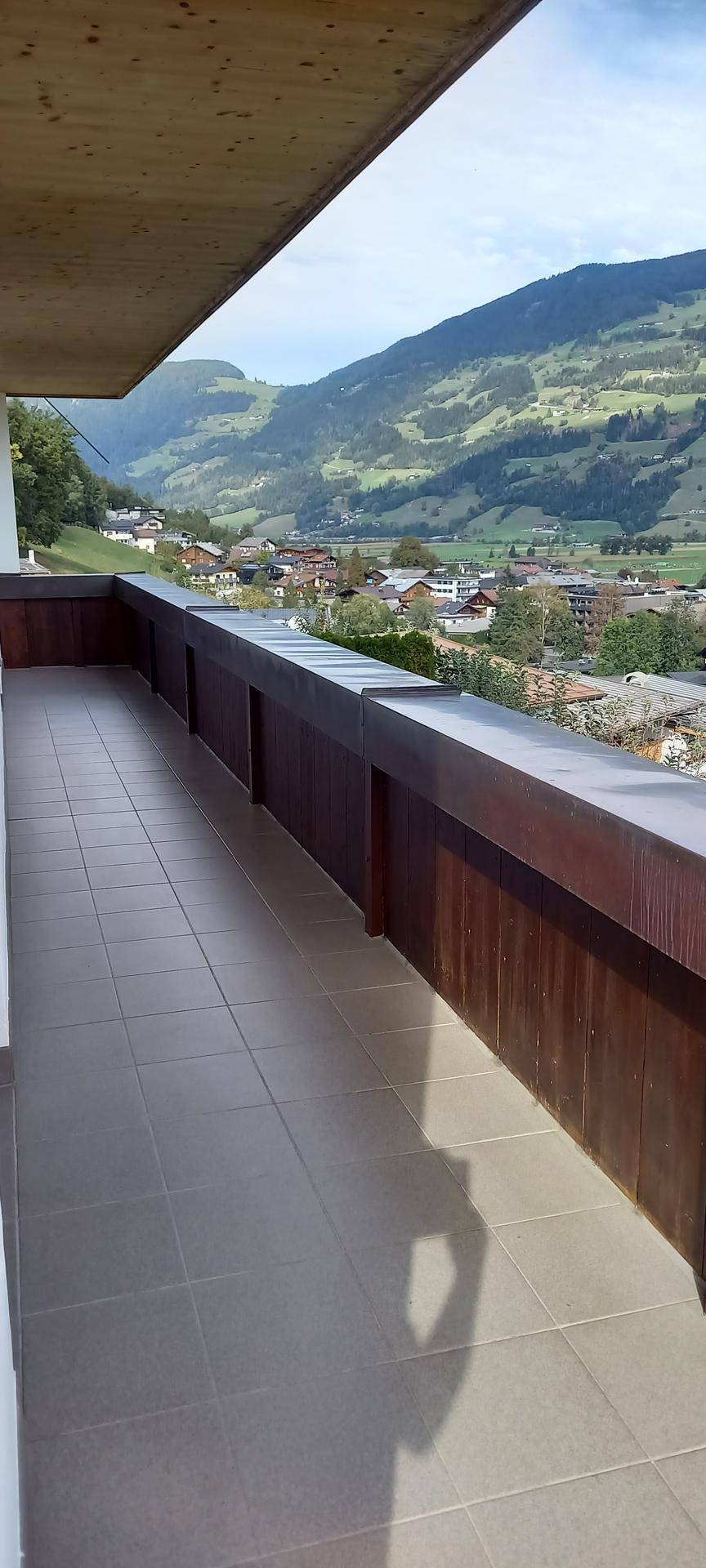 Langer Balkon mit Fliesenboden und Holzgeländer, der einen weiten Blick auf die Berge und das Tal bietet.