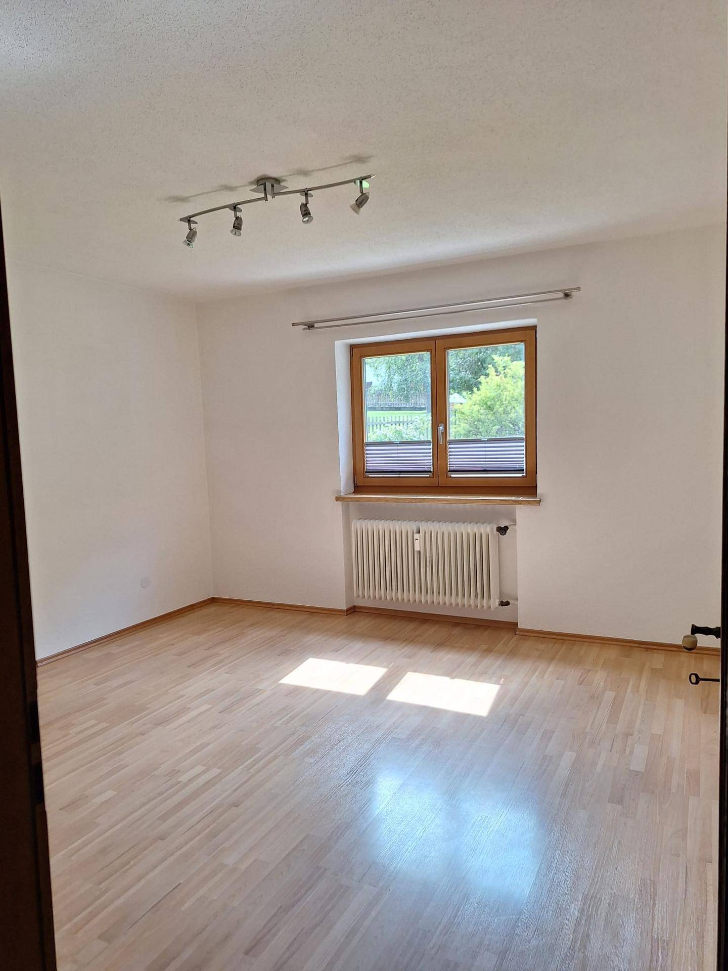 Lichtdurchfluteter Raum mit Laminatboden, Heizkörper und Blick ins Grüne durch das Fenster.
