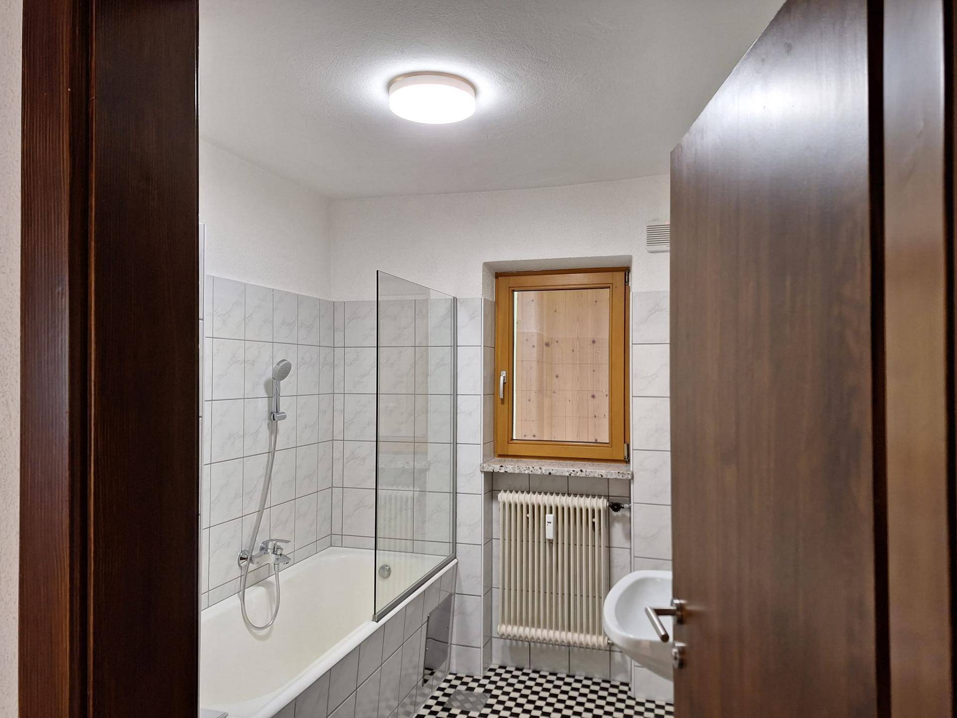 Badezimmer mit Badewanne, Duschabtrennung, Fenster und schwarz-weiß kariertem Fliesenboden.