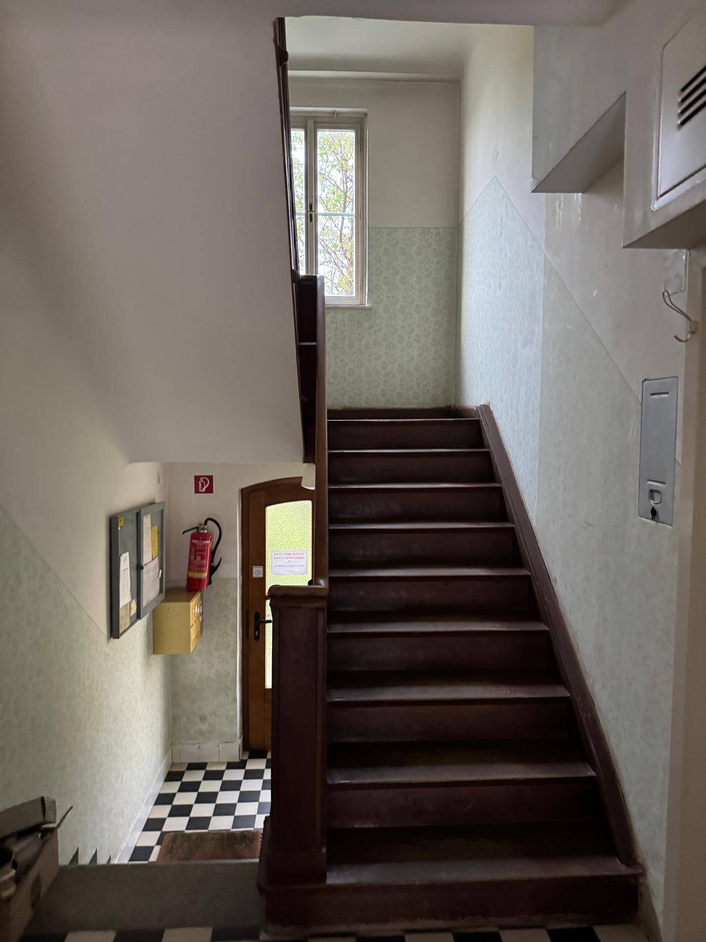 Treppenhaus mit Holztreppe und Schachbrettmuster-Fliesenboden, führt zu den oberen Etagen.
