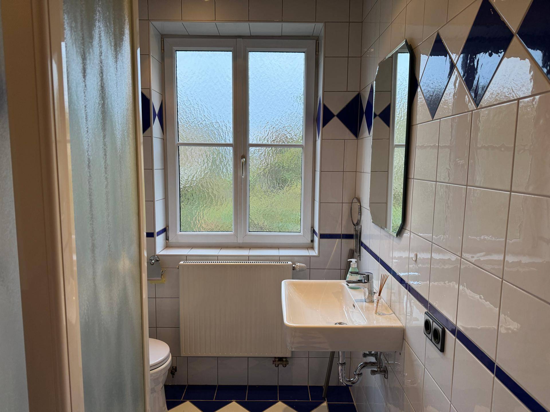 Badezimmer mit Waschbecken, Toilette und Fenster, ausgestattet mit weißen und blauen Fliesen.