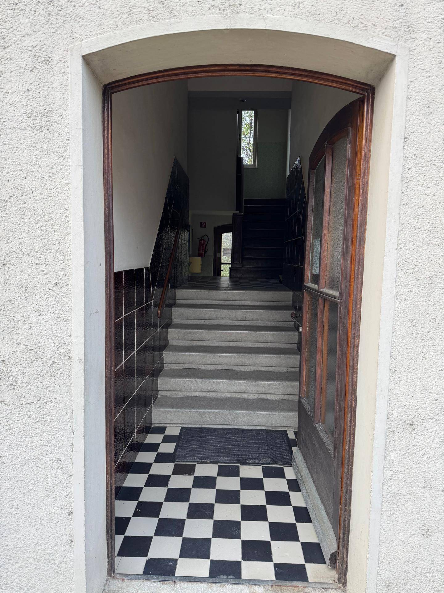 Eingangsbereich eines Gebäudes mit Schachbrettmuster-Fliesenboden und Treppenhaus.