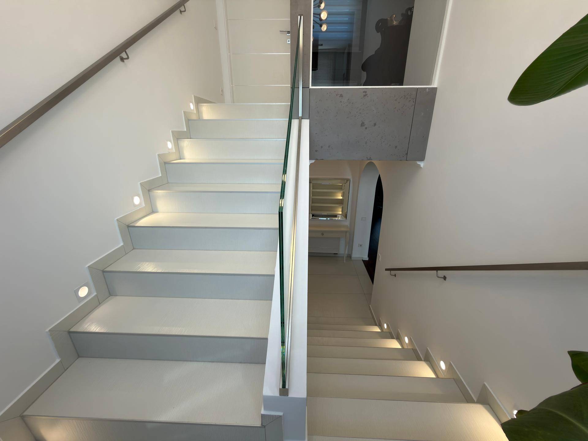 Moderne Treppe mit Glasgeländer und indirekter Beleuchtung, die zu den unteren Ebenen führt.