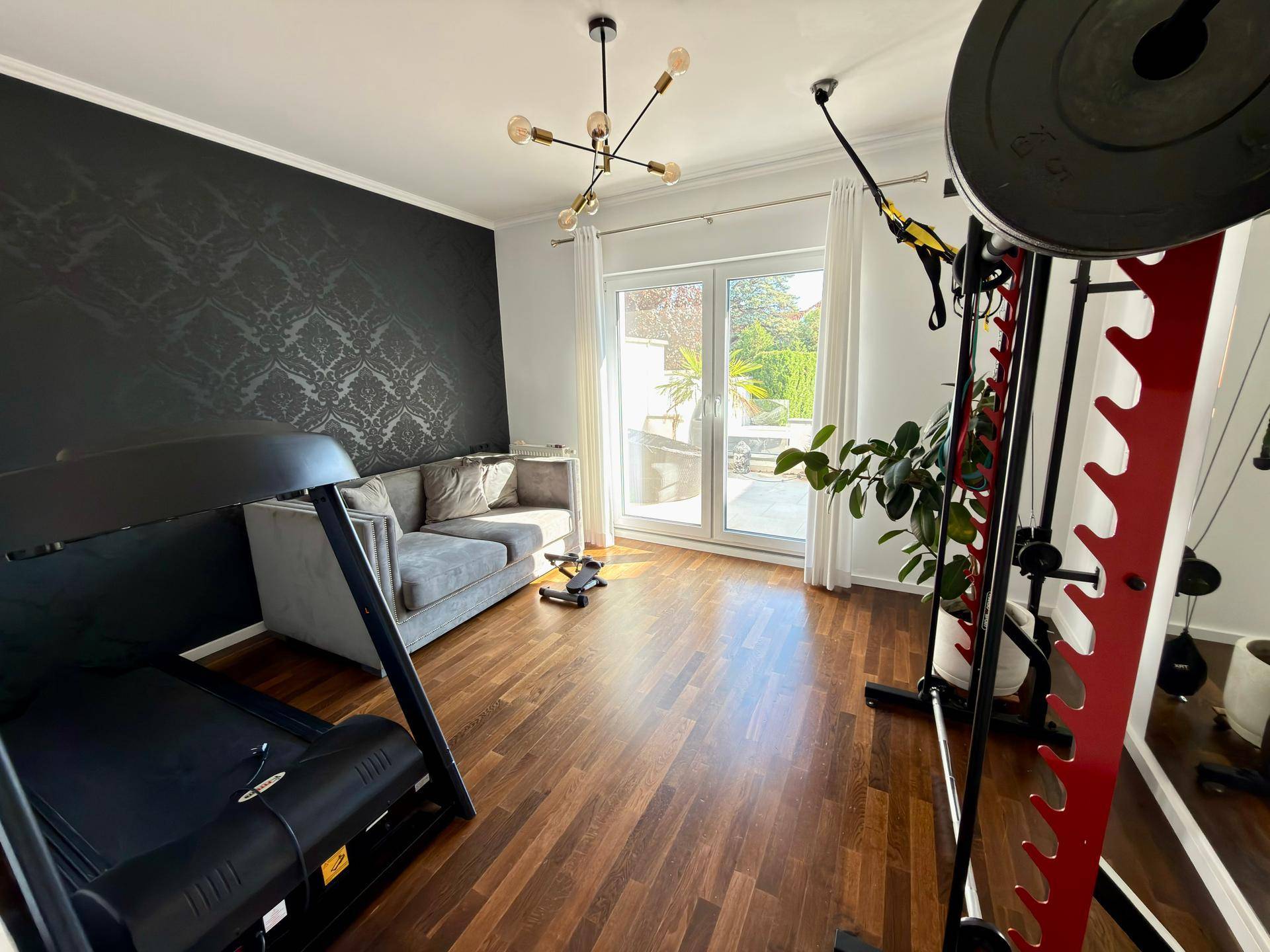 Fitnessraum mit Laufband, Gewichten und Zugang zu einem sonnigen Außenbereich.