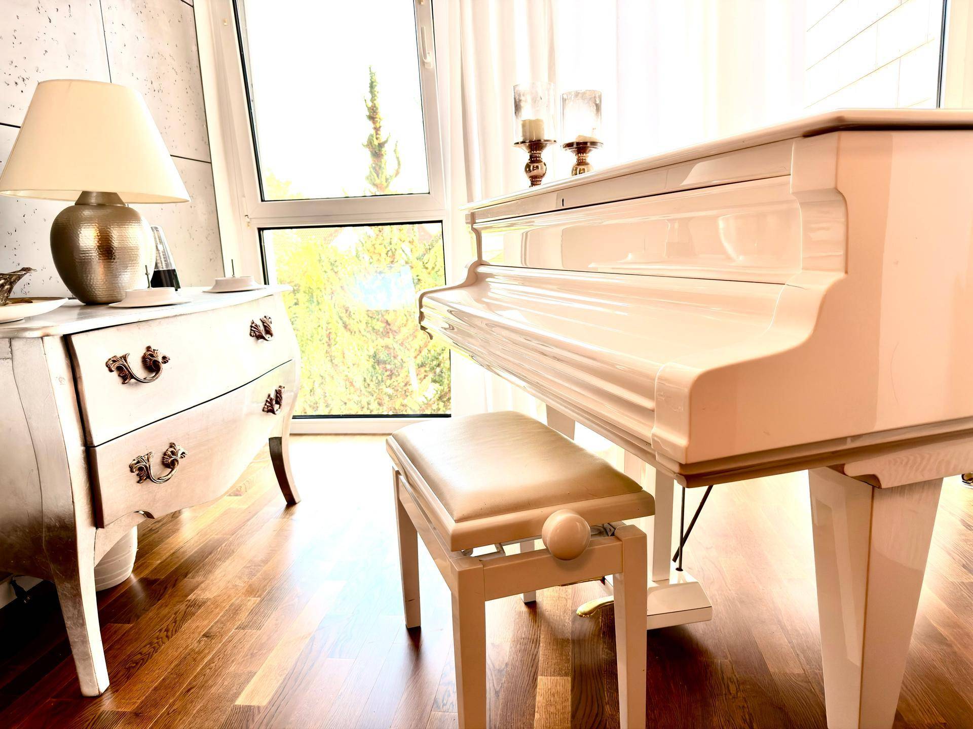 Elegantes weißes Klavier und antike Kommode in einem hellen Raum mit Holzboden und großen Fenstern.