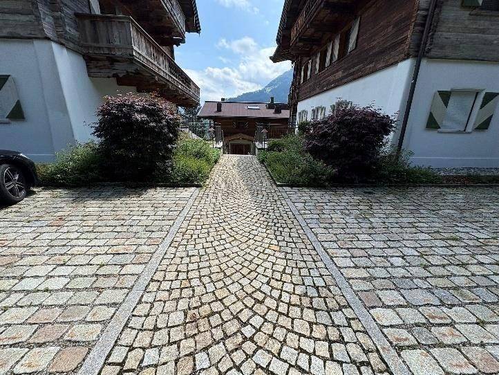 Kopfsteinpflaster-Einfahrt zwischen zwei traditionellen Chalet-Gebäuden mit Bergblick im Hintergrund.