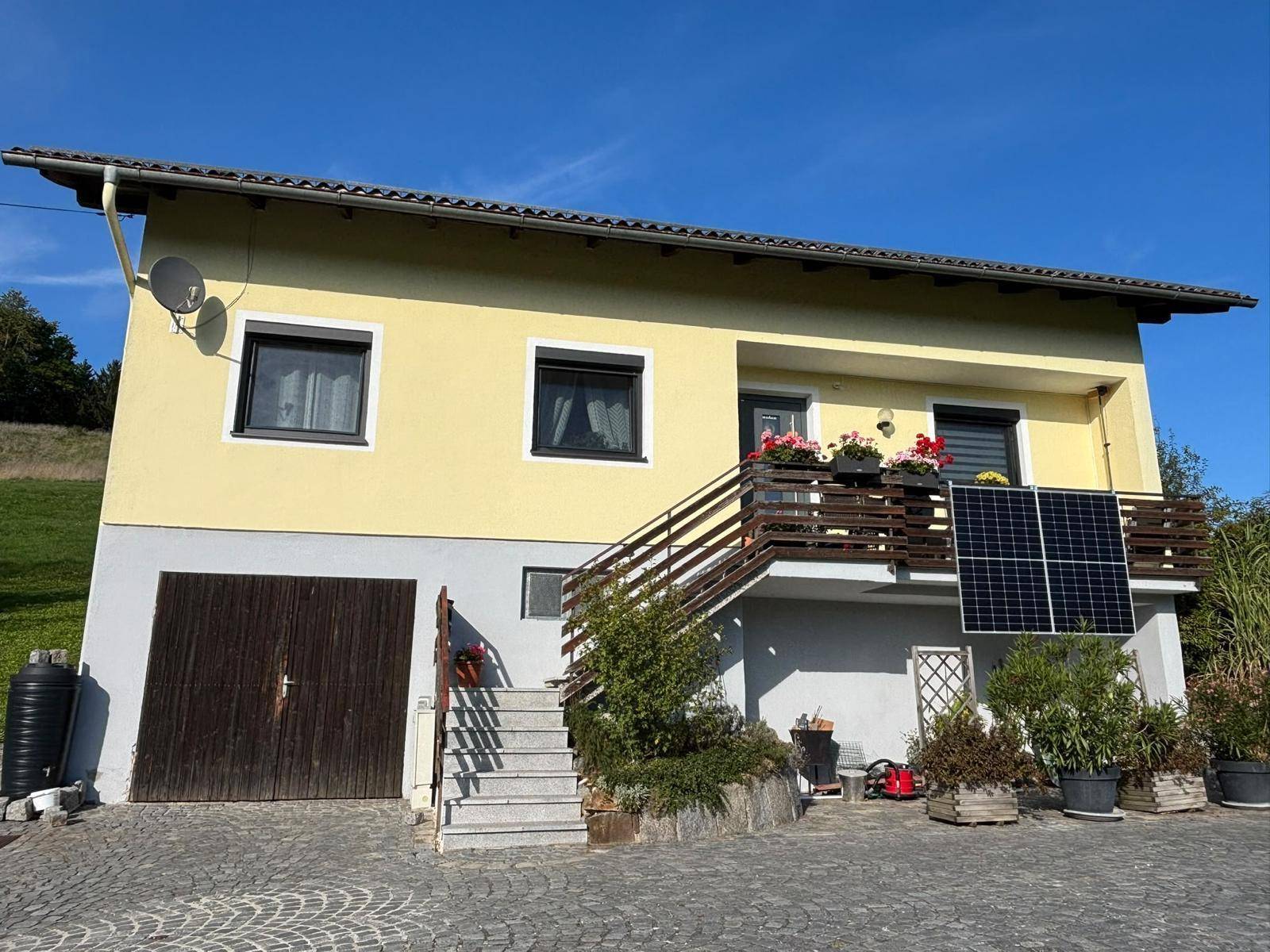 Vorderansicht des Hauses mit gelber Fassade, Garage, Außentreppe und Solarmodulen am Balkon.
