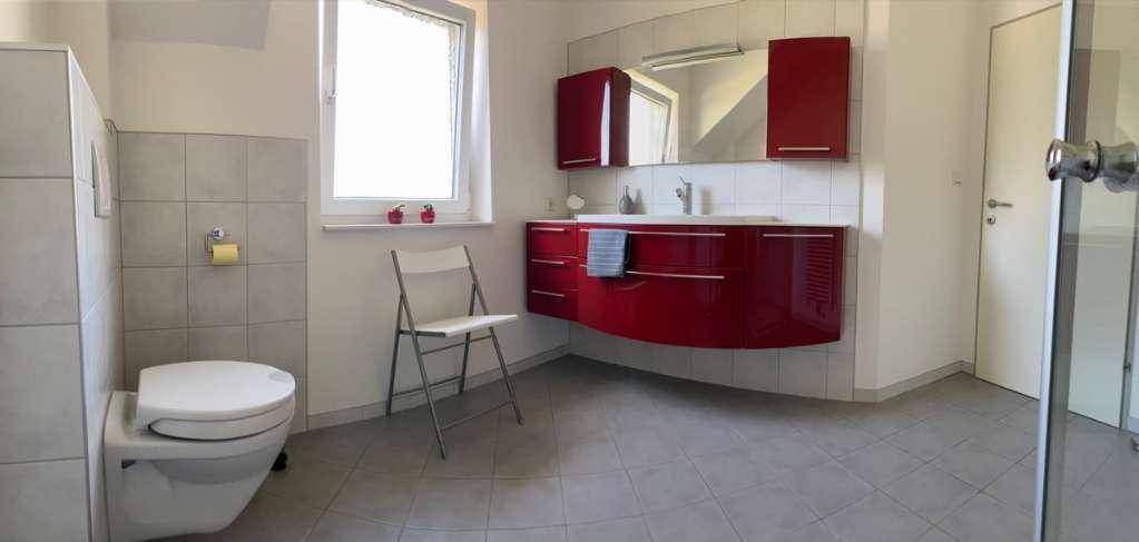 Helles Badezimmer mit roten Badmöbeln, Toilette und Zugang zur Dusche.