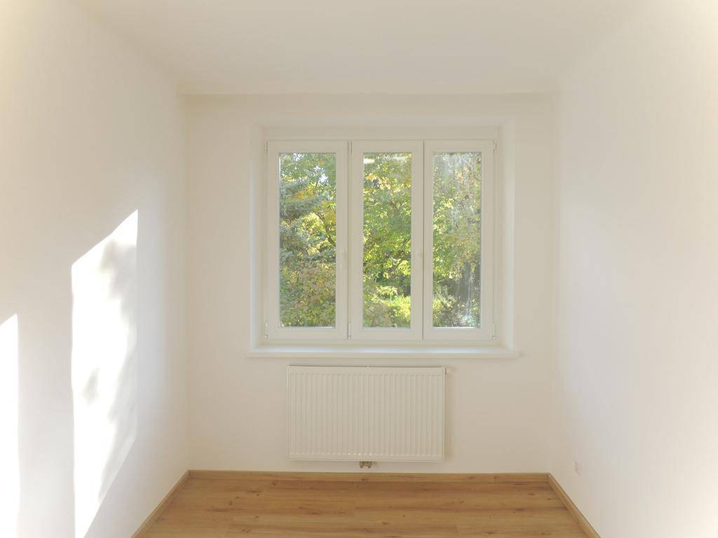 Heller Raum mit Laminatboden und großem Fenster, das viel Tageslicht und Grünblick bietet.