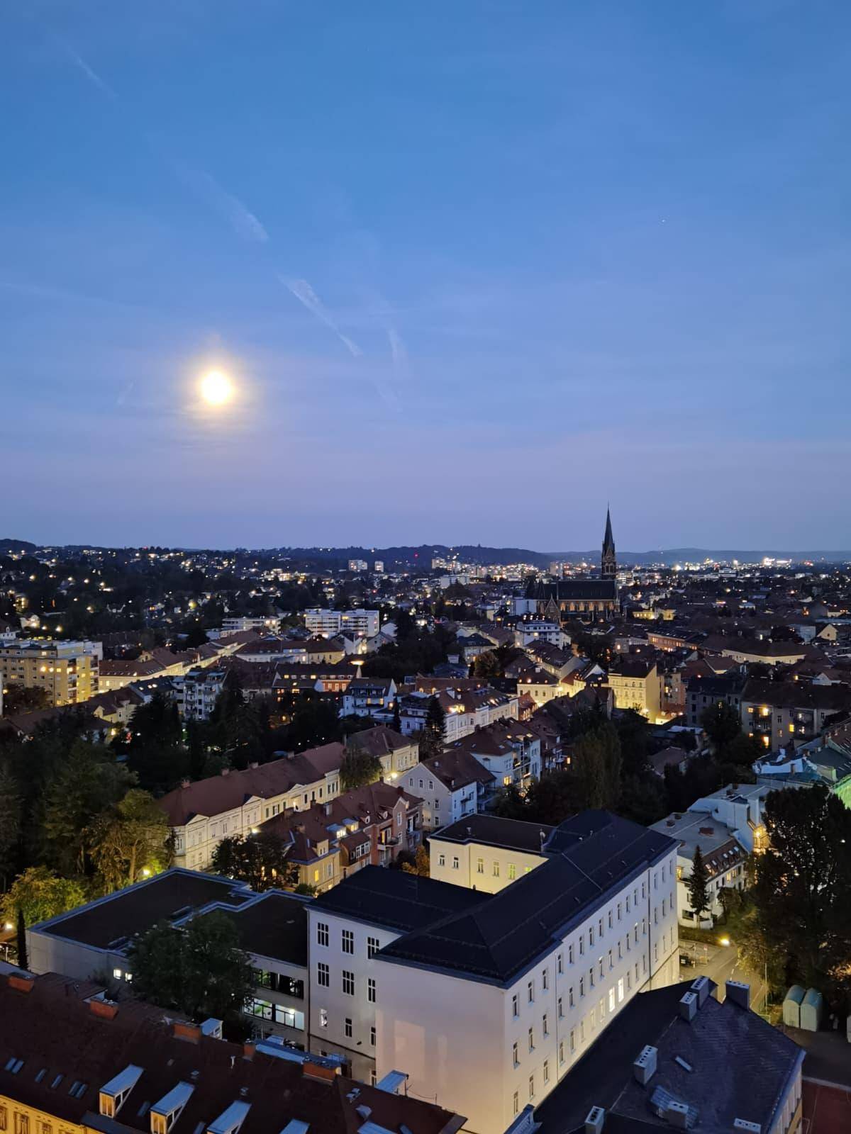 Nachtaufnahme der Stadt mit leuchtenden Lichtern und Vollmond am dunklen Himmel.