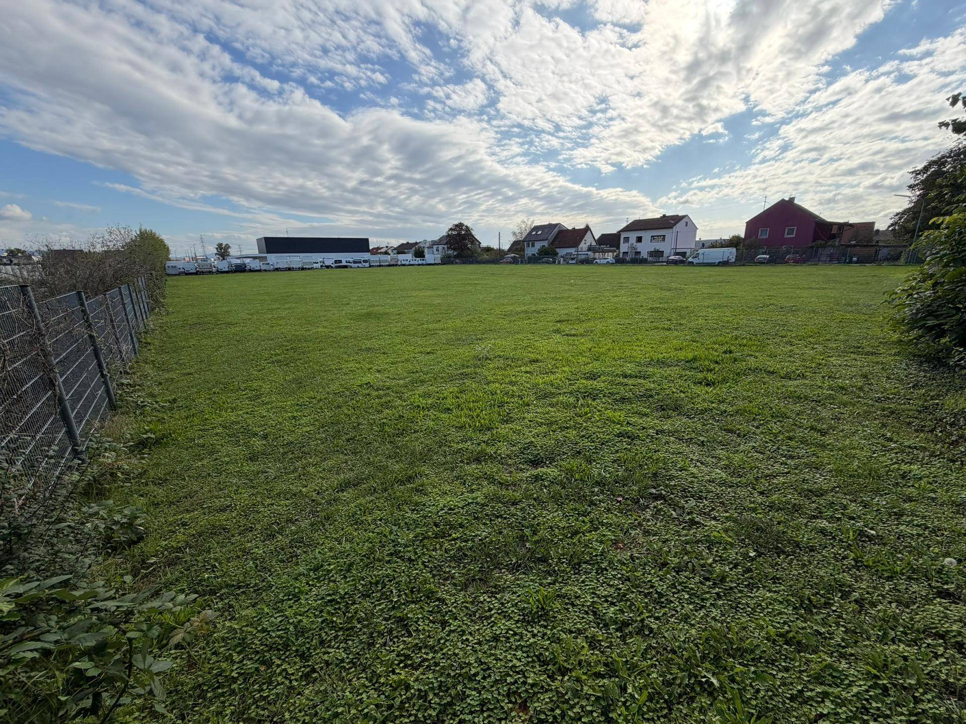 Weitläufiges grünes Feld mit Häusern und Industriegebäuden im Hintergrund unter bewölktem Himmel.