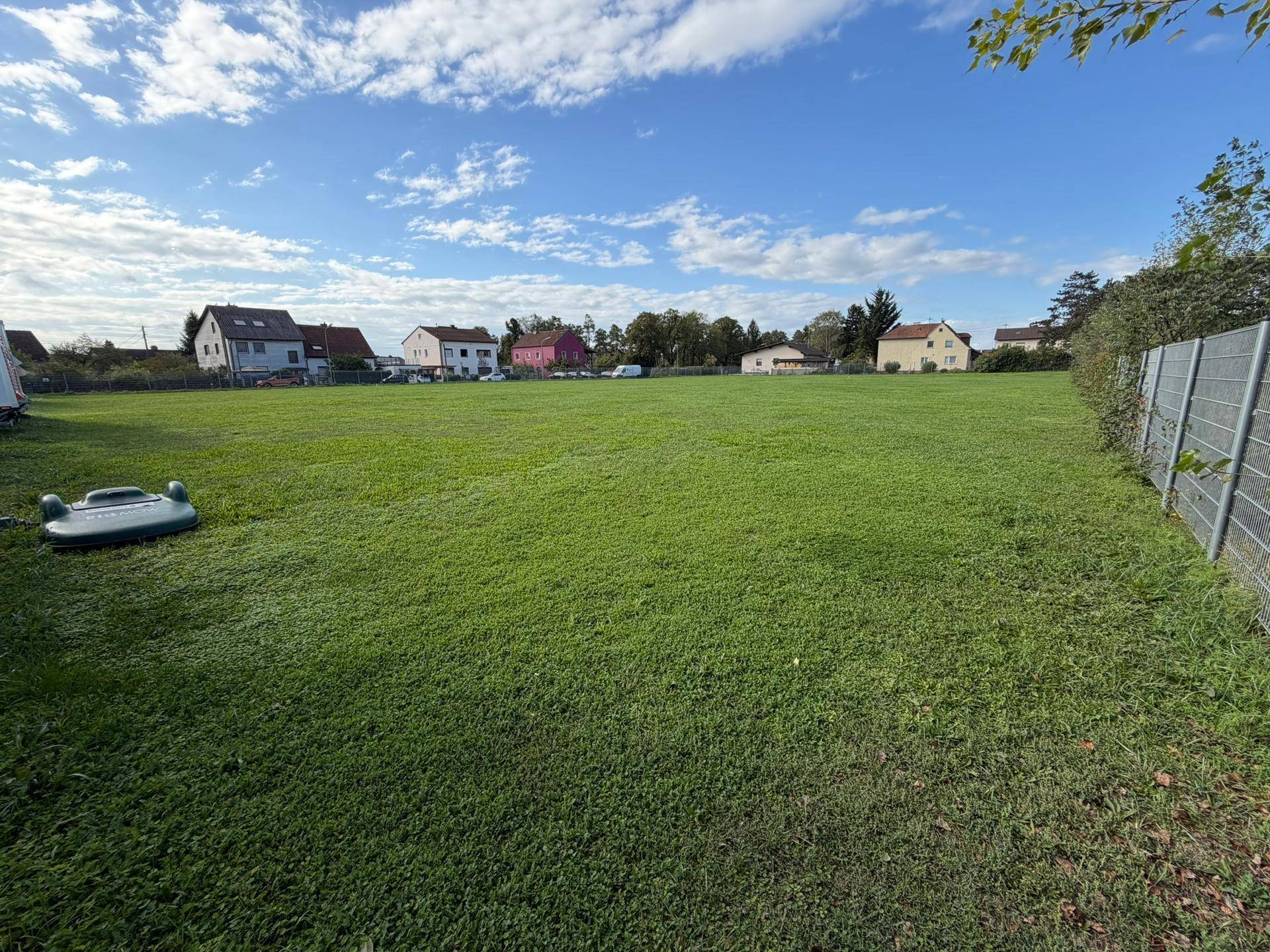 Offenes Wiesengrundstück mit Wohngebäuden im Hintergrund und einem Zaun auf der rechten Seite.