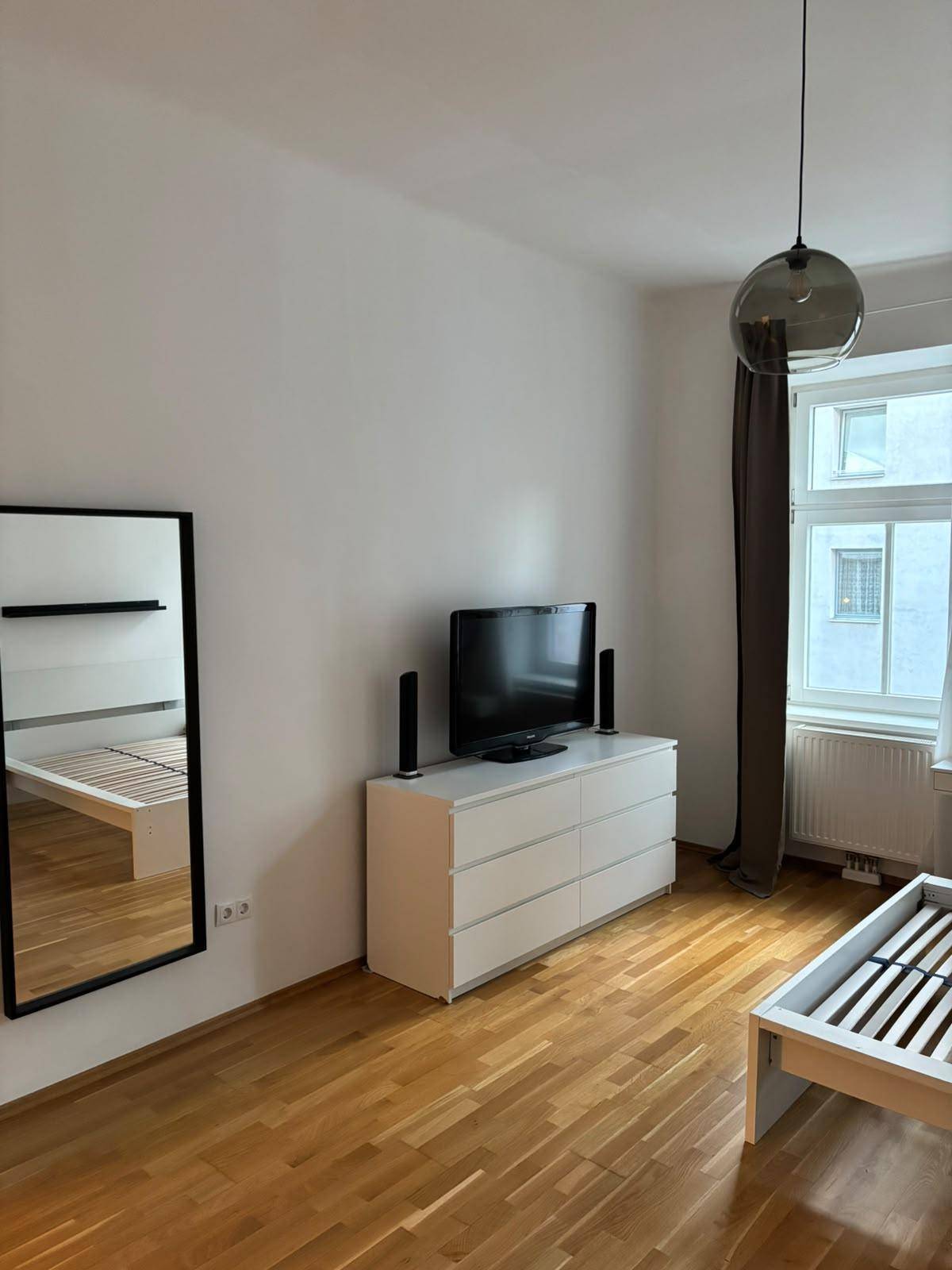 Schlafzimmer mit Fernseher auf einer Kommode, einem Spiegel und hellem Holzboden.