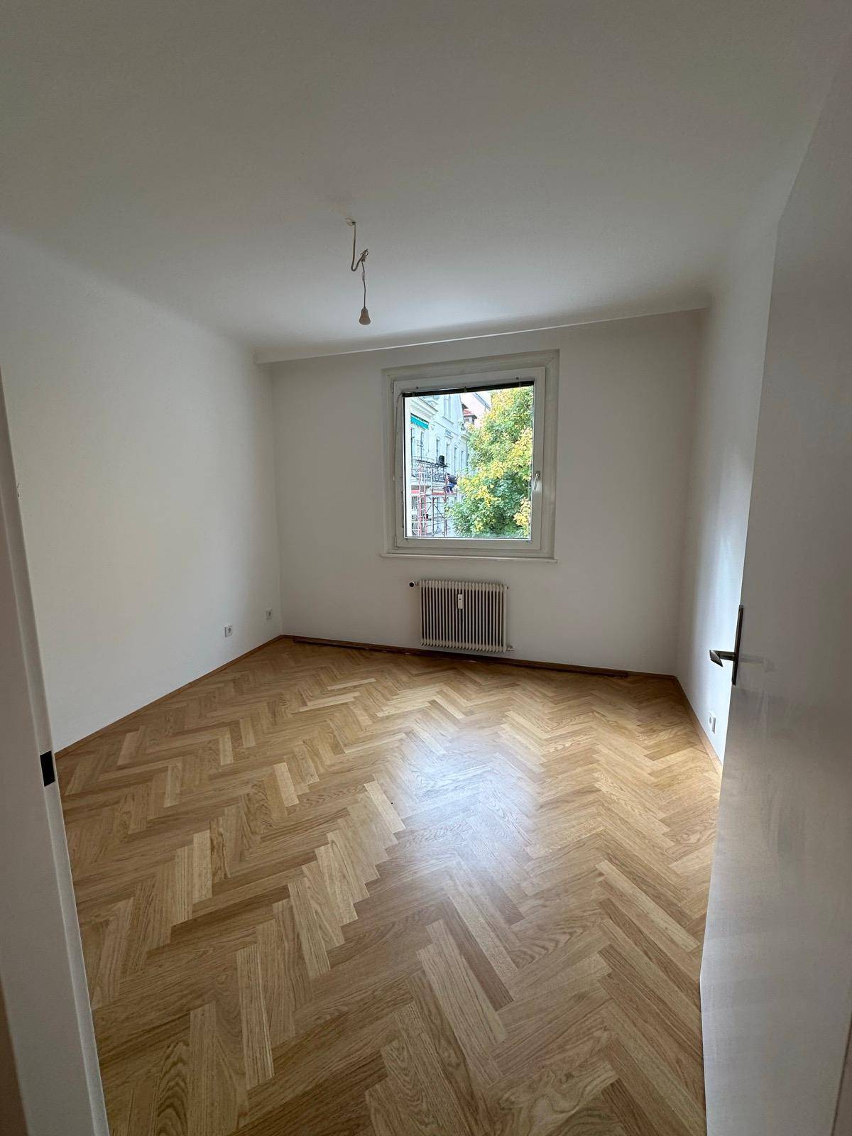 Helles Zimmer mit klassischem Fischgrätparkett und einem Fenster, das den Blick auf die Straße freigibt.