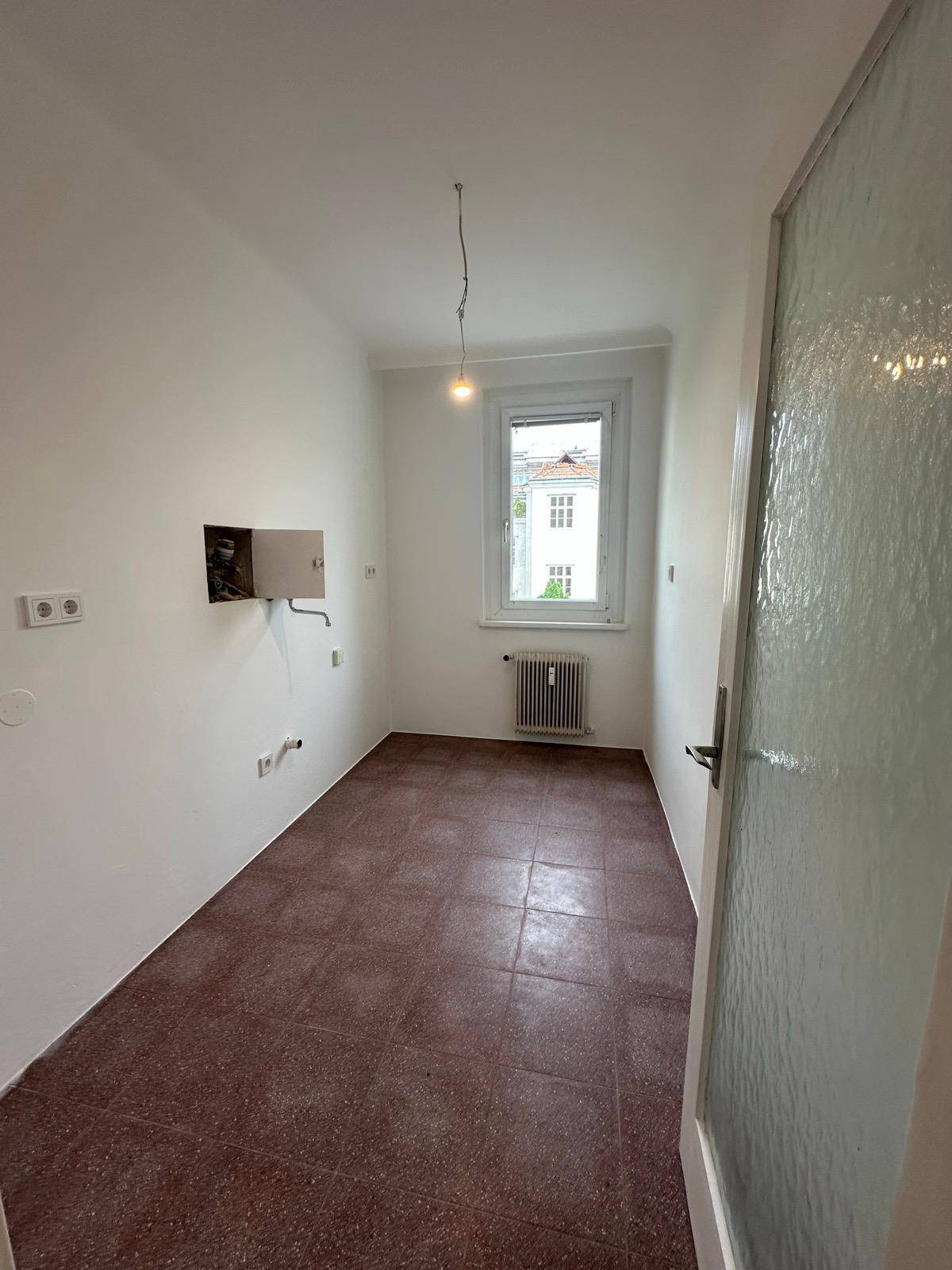 Leerer Raum mit braunen Fliesen, einem Fenster und Heizkörper, bereit für die individuelle Gestaltung und Nutzung.