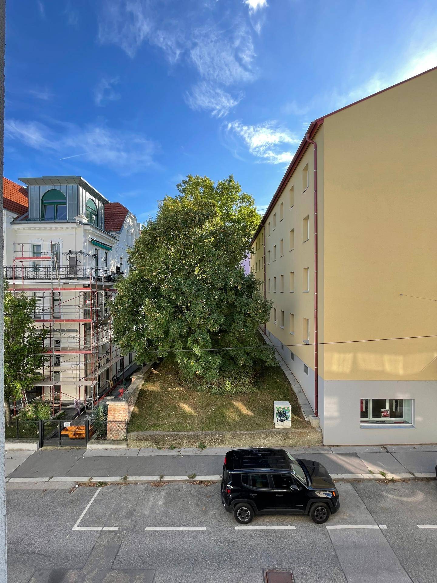 Blick aus dem Fenster auf eine Straße mit parkenden Autos und umliegenden Gebäuden, eines davon eingerüstet.