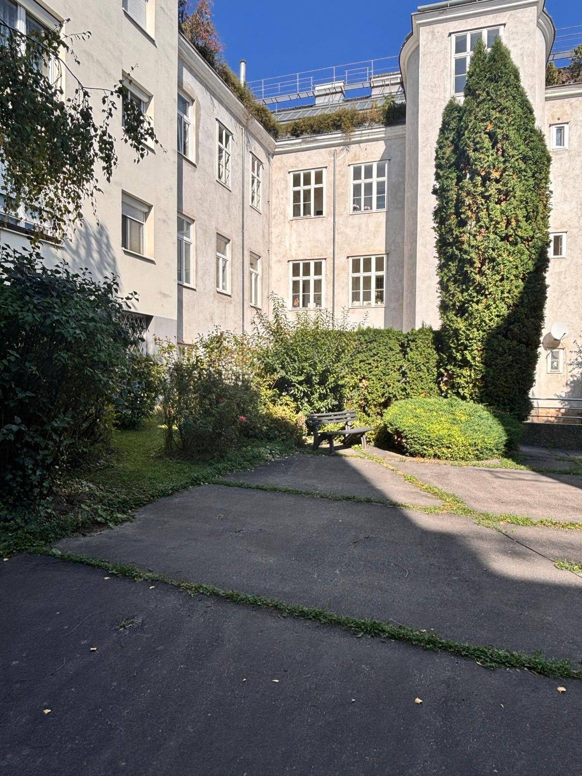 Innenhof mit einer Holzbank, umgeben von Grünflächen und hohen Bäumen, zwischen den Gebäuden gelegen.