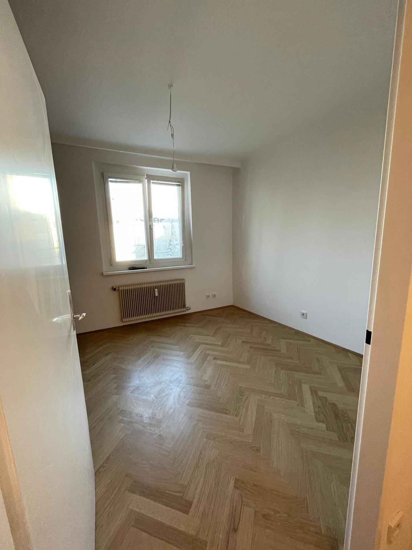 Helles Zimmer mit elegantem Fischgrätparkettboden und einem großen Fenster, das viel Tageslicht hereinlässt.