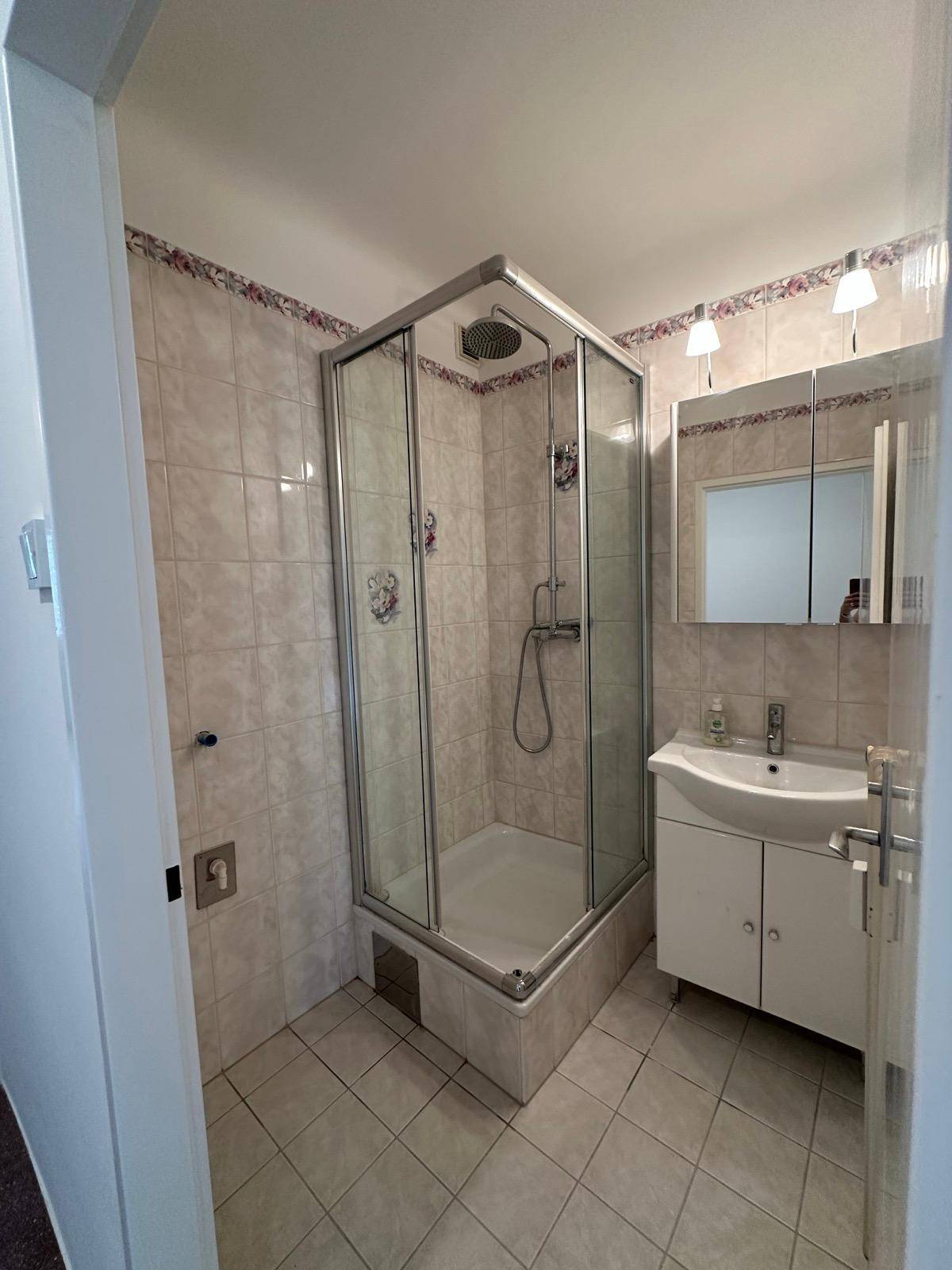 Badezimmer mit Eckduschkabine, Waschbecken mit Unterschrank und hellen Fliesen an Wänden und Boden.