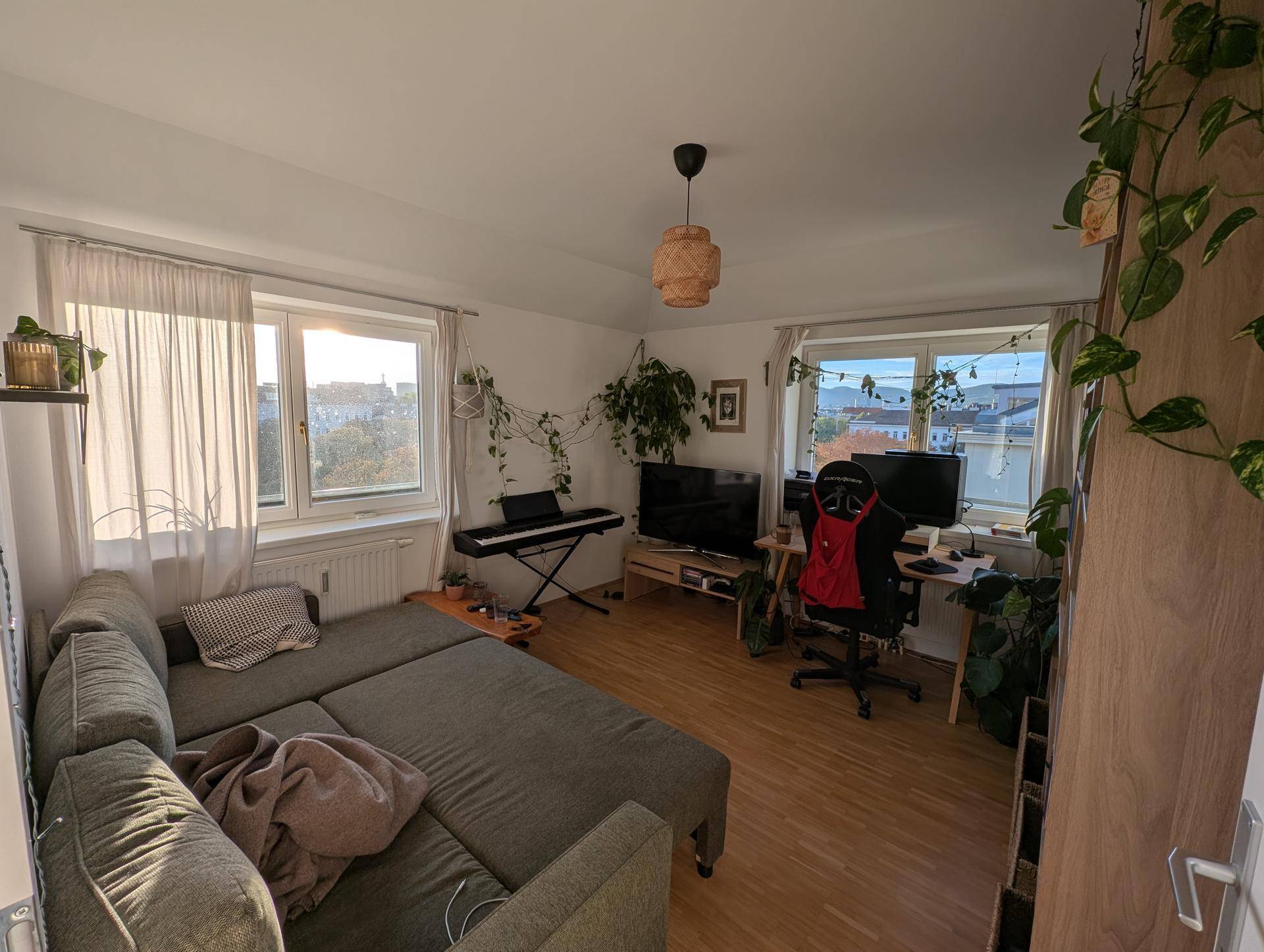 Gemütliches Wohnzimmer mit Sofa, Schreibtisch und vielen Pflanzen, hell durch große Fenster.
