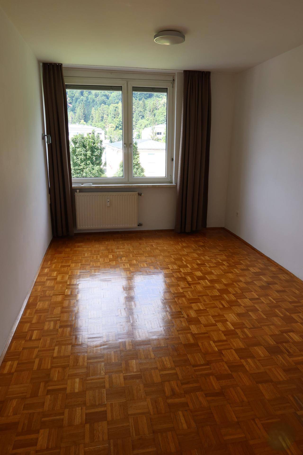 Leeres Schlafzimmer mit Parkettboden und großem Fenster mit Blick ins Grüne.