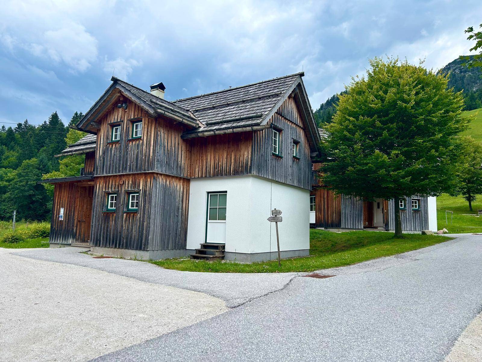 Traditionelles Holzhaus im Chalet-Stil mit weißem Sockel und grüner Umgebung vor Bergkulisse.