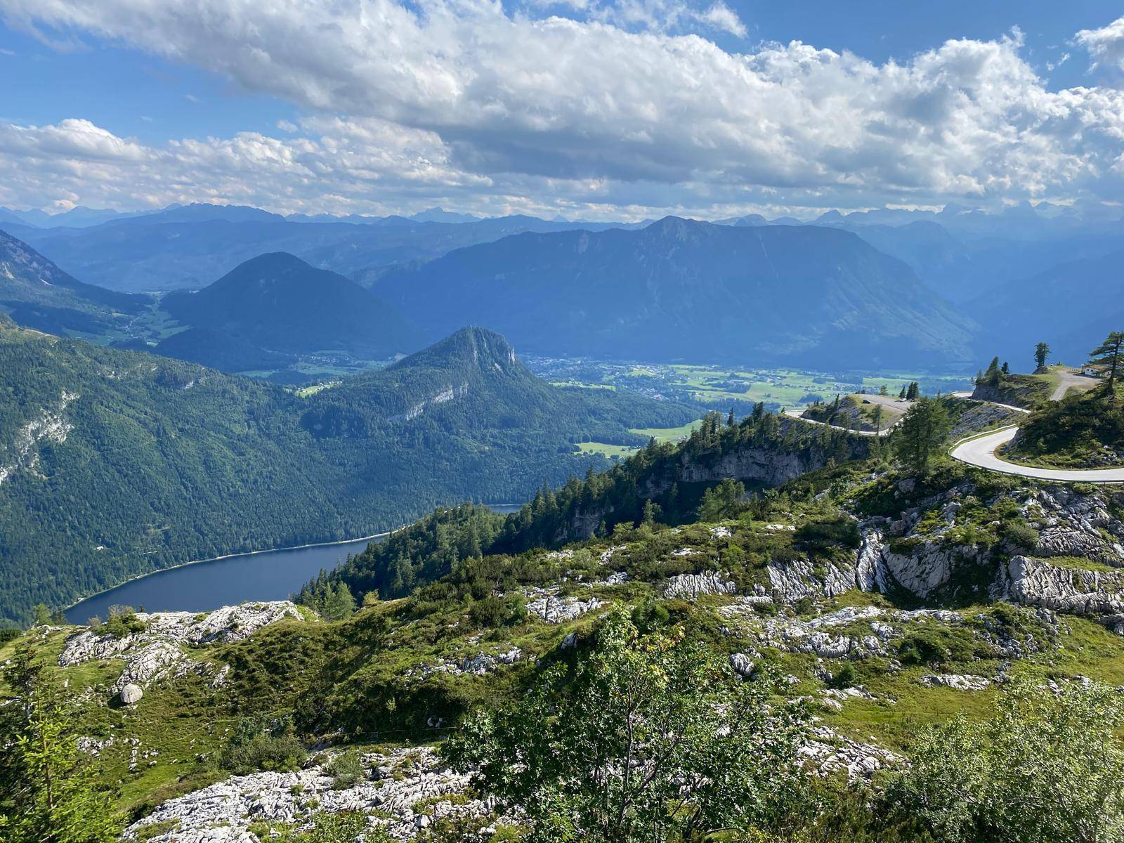 Atemberaubende Panoramaaussicht auf eine Berglandschaft mit See, grünen Tälern und kurvenreicher Straße.