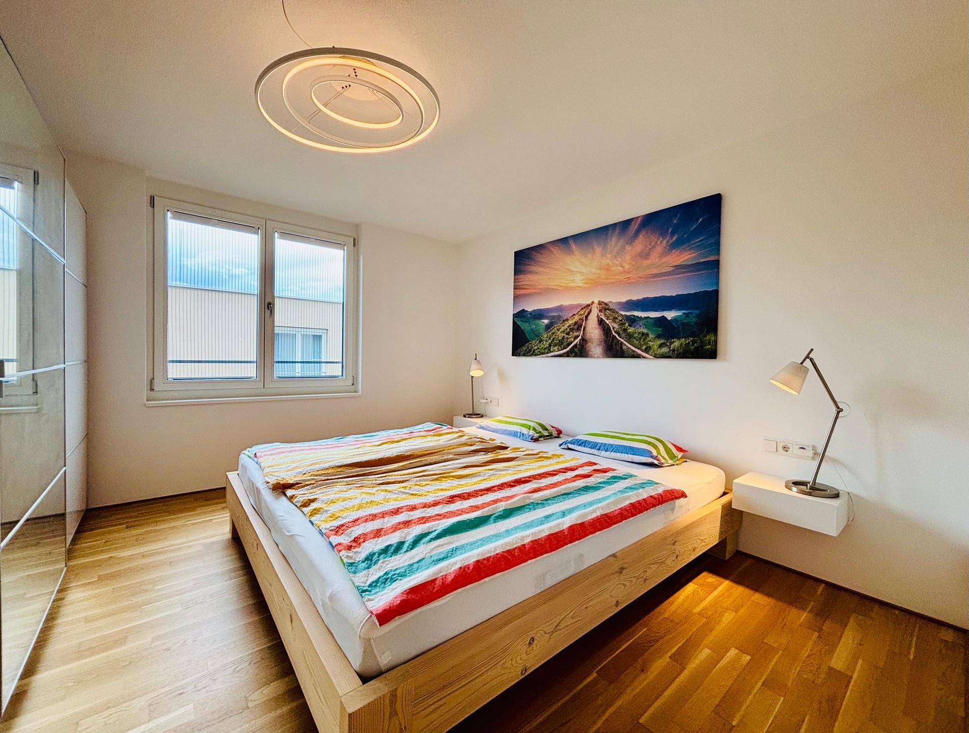 Schlafzimmer mit Holzbett, farbenfroher Bettwäsche und großem Fenster für viel Licht.
