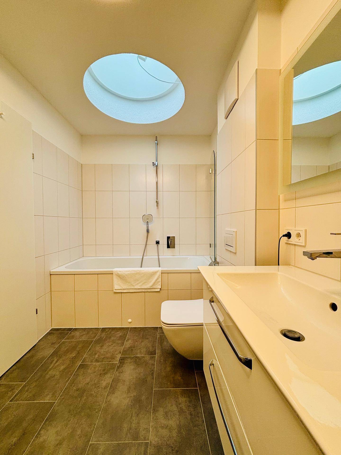 Helles Badezimmer mit Badewanne, Toilette, Waschbecken und Oberlicht für Tageslicht.