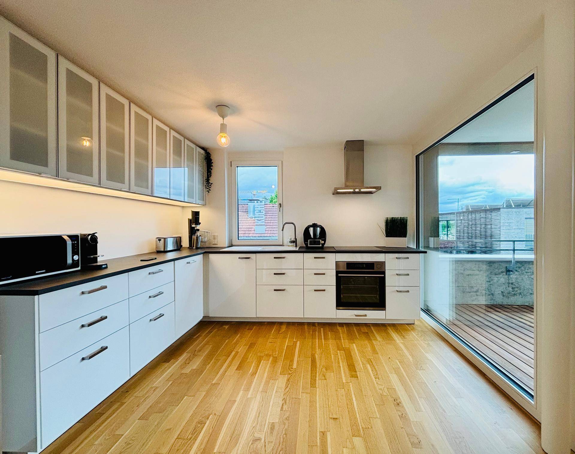 Moderne Küche mit weißen Schränken, dunkler Arbeitsplatte und Zugang zum Balkon.