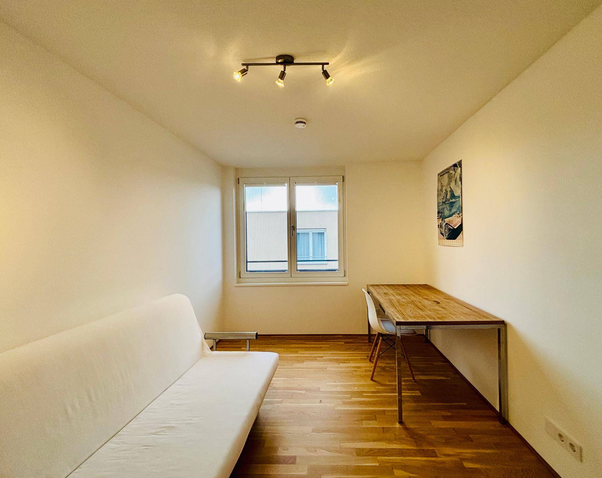 Helles Zimmer mit Sofa, Schreibtisch und Fenster, ideal als Gäste- oder Arbeitszimmer.