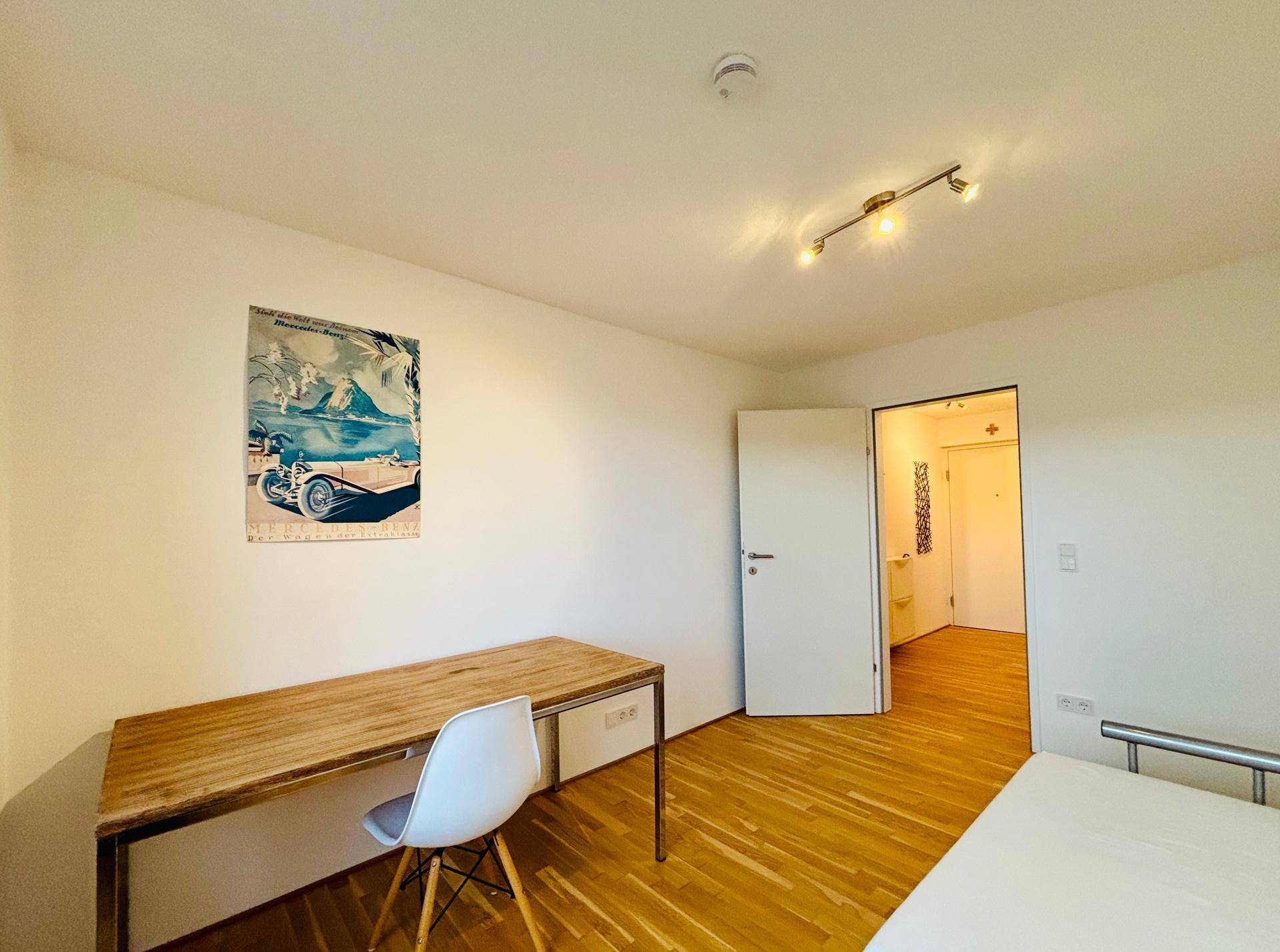 Helles Zimmer mit Holzboden, Schreibtisch und Zugang zu einem Badezimmer.
