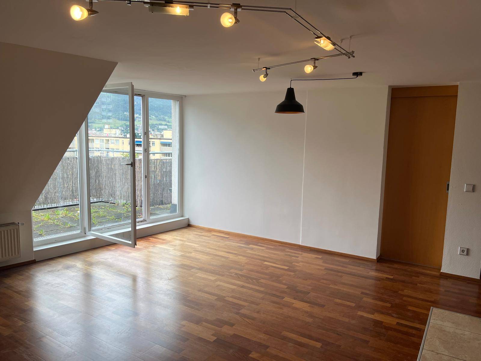 Heller Wohnbereich mit Holzboden, Dachschräge und großem Fenster mit Zugang zum Balkon.