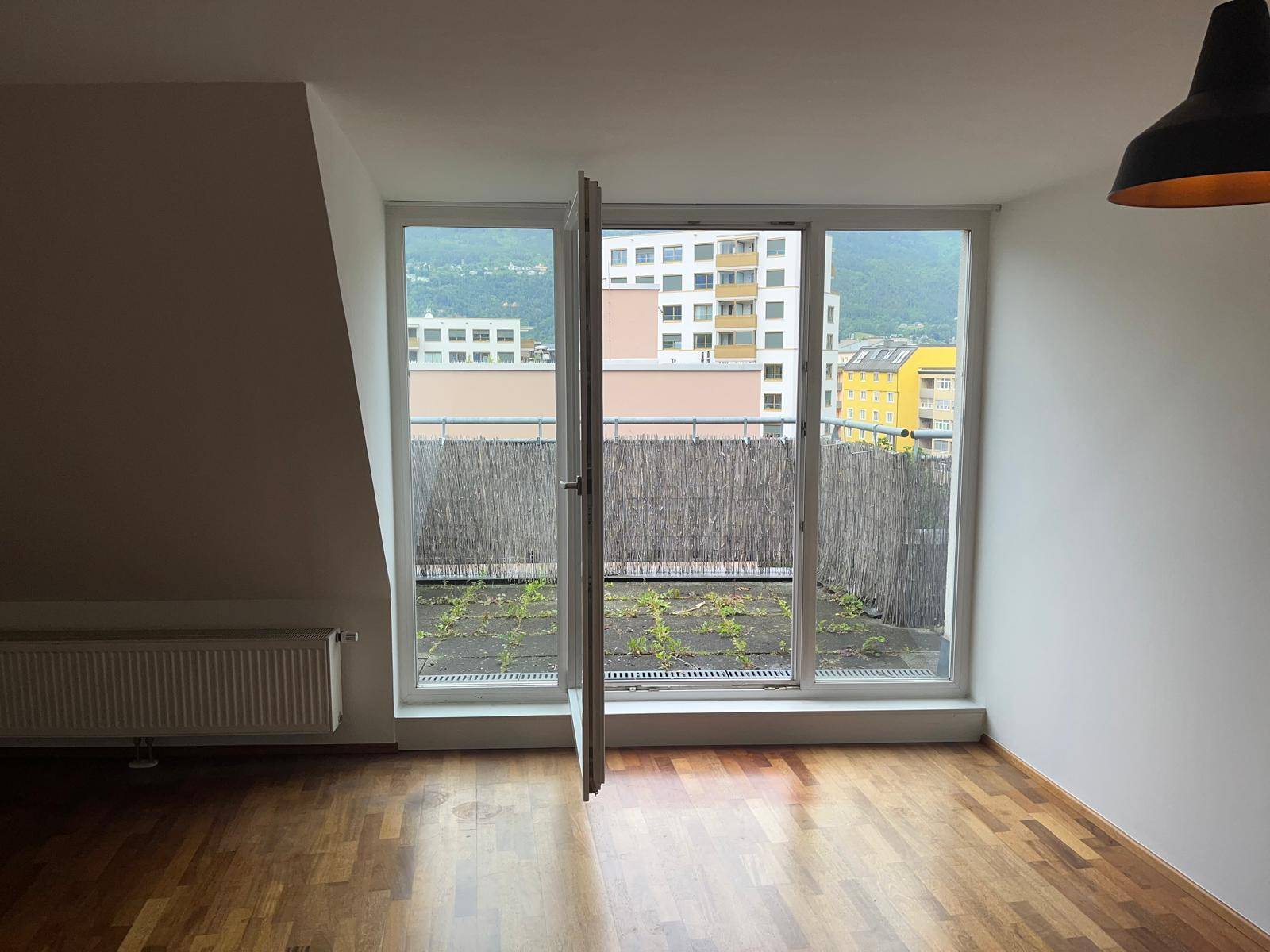 Geräumiger Wohnbereich mit Holzboden und großem Fenster, das Zugang zu einem Balkon bietet.